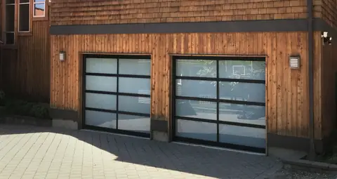 Two single full-view garage doors with black frames and white laminated glass, set in cedar board siding above a paver driveway