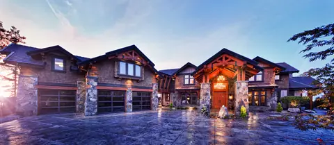 Three full-view garage doors with black frames and dark glass between stone pillars on a large timber and shingle house at dusk