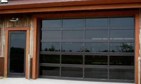 Full-view garage door, 4 lites by 5, dark bronze frame and gray tinted glass, on a barn-board building by a glass door