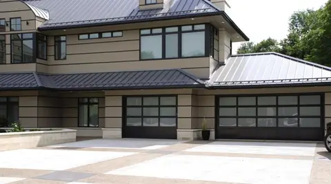Two full-view garage doors with black frames, patterned frosted glass above and a solid black bottom section, on a tan banded house