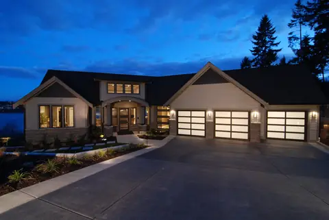 Three single full-view garage doors with dark bronze frames and white laminated glass, lit from inside at dusk, on a beige house