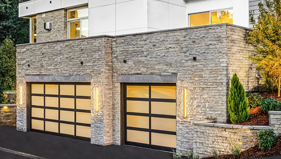 A double and a single full-view garage door with black frames and white laminated glass, lit from inside, in stacked stone