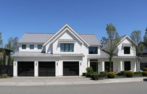 Three single full-view garage doors with black frames and black laminated glass on a white board-and-batten house with a metal roof