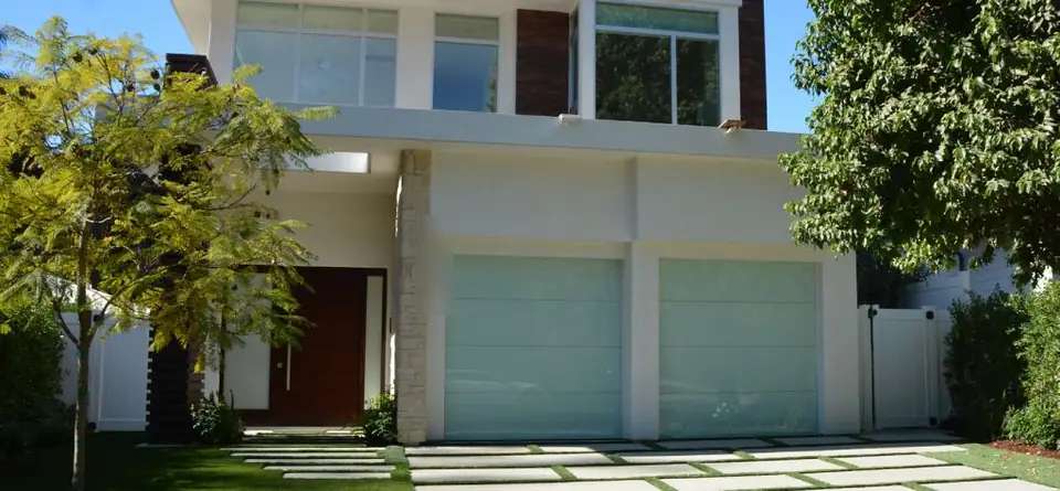 Two single frameless full-view garage doors in pale frosted glass on a white stucco house, with a paver and grass driveway