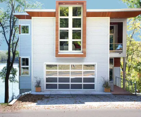 White-framed full-view aluminum garage door with mirror glass, four panes across, on a white corrugated metal house by a lake.