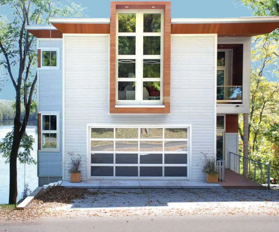 White-framed full-view aluminum garage door with mirror glass, four panes across, on a white corrugated metal house by a lake.