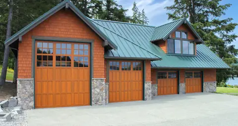 Four wood-look carriage garage doors with divided-lite windows, one extra tall, on a cedar shingle shop with a green metal roof.