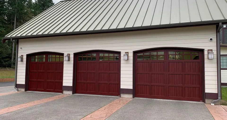 Three arched-top wood-look carriage garage doors in dark red with V-groove panels and divided lites, on a cream sided garage.