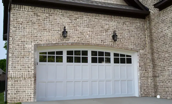 White arched-top carriage-style double garage door with a row of divided-lite windows, set in a light gray brick wall.