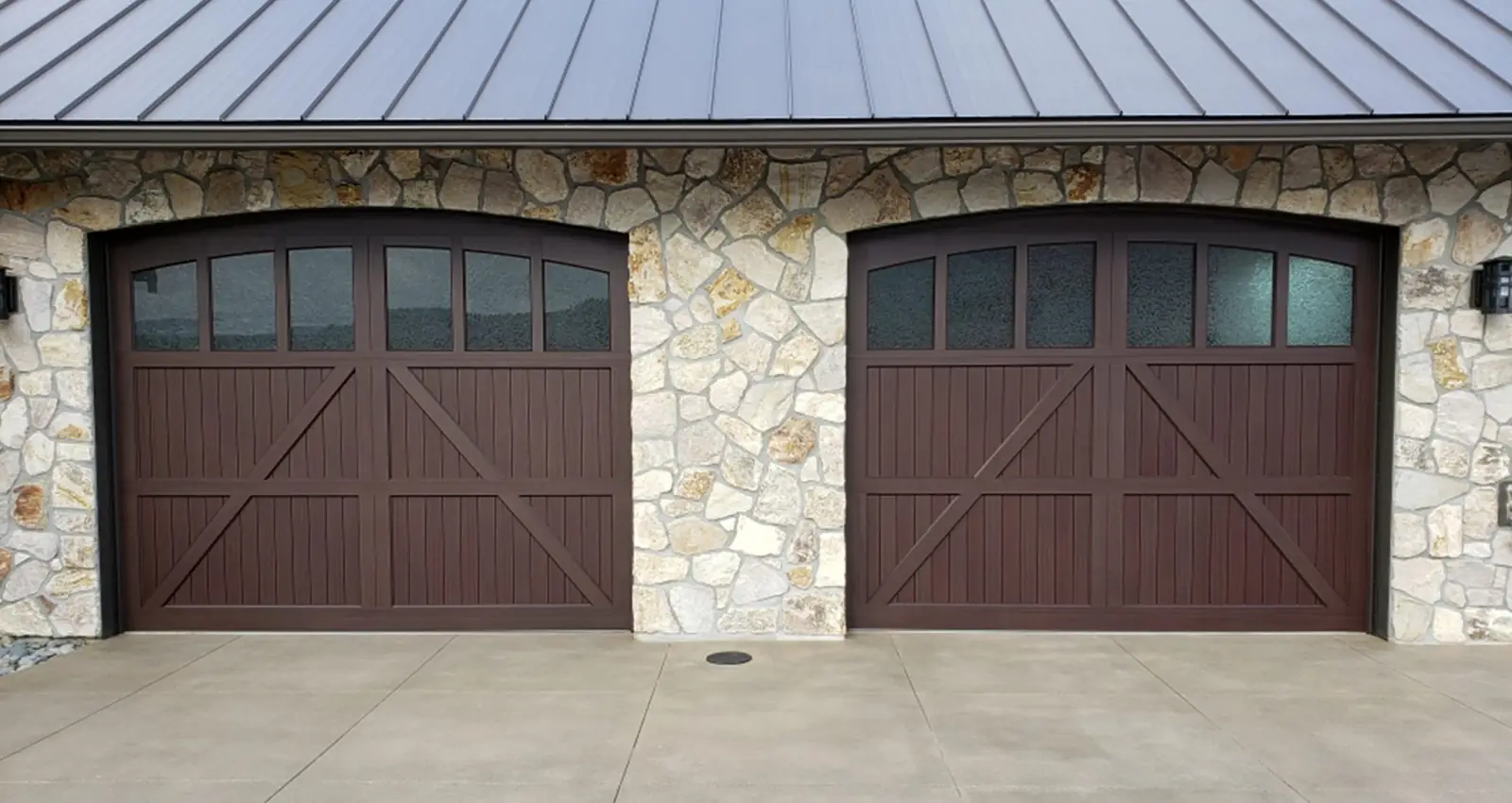 Two arched-top dark brown garage doors with V-groove boards, X bracing and a row of glass lites, set in a fieldstone wall.