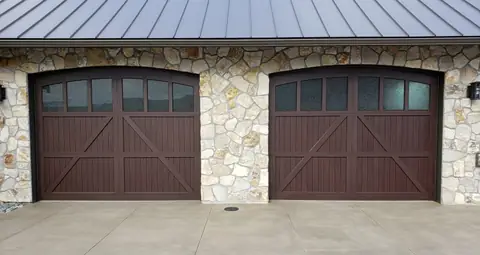 Two arched-top dark brown garage doors with V-groove boards, X bracing and a row of glass lites, set in a fieldstone wall.