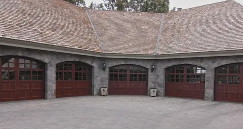 Five arched-top carriage garage doors in a dark red-brown wood finish with divided lites, set in gray stone arches.
