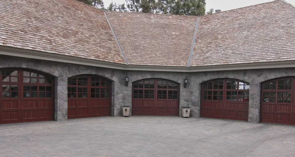 Five arched-top carriage garage doors in a dark red-brown wood finish with divided lites, set in gray stone arches.