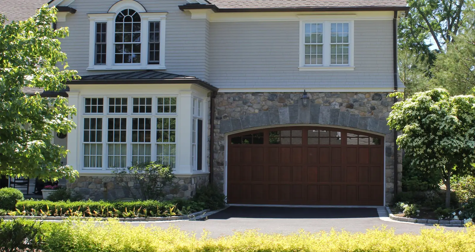 Double-width arched-top carriage garage door in a dark brown wood finish with a row of top lites, set in a fieldstone wall.