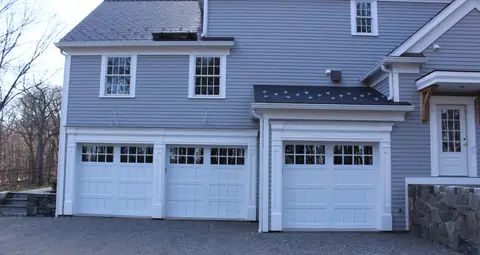 Three white garage doors with small recessed panels and divided-lite top rows, in white trim on a gray clapboard house.