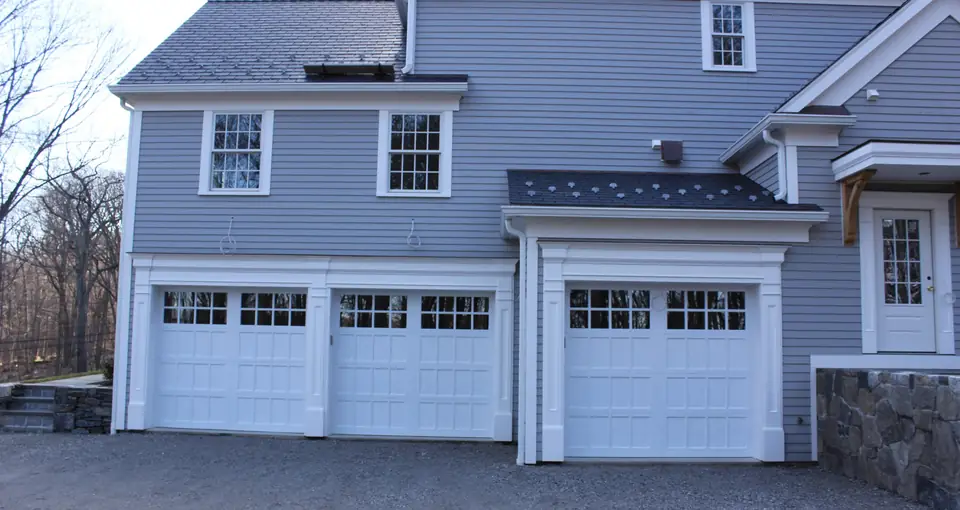 Three white garage doors with small recessed panels and divided-lite top rows, in white trim on a gray clapboard house.