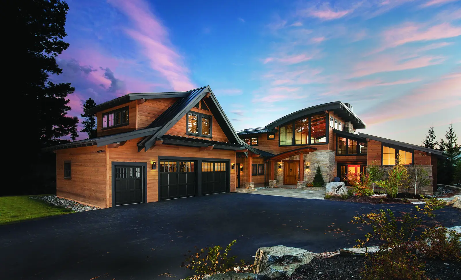 Three black carriage-style garage doors with divided-lite top rows on a cedar-clad garage wing, photographed at dusk.