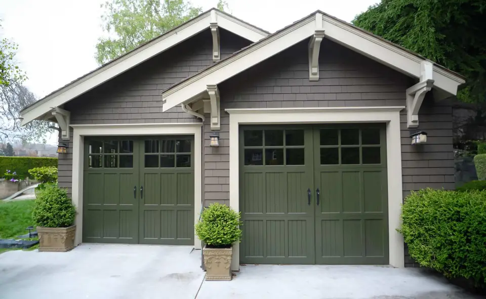 Two olive green carriage-style garage doors with V-groove panels, clear glass top lites and black handles, on a shingled garage.