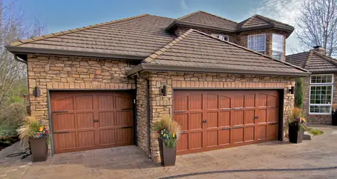 Two carriage-style garage doors in a mahogany wood-look finish with arched top panels and black strap hinges, on a stone house.