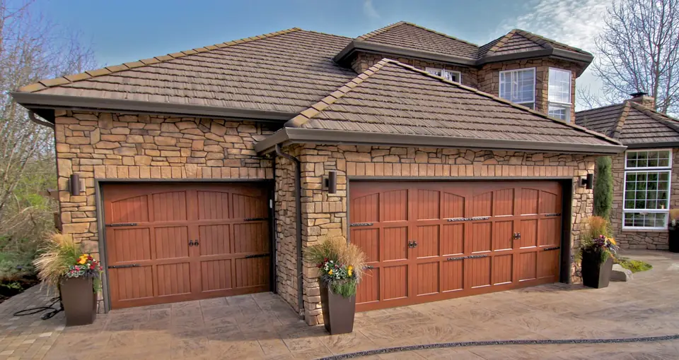 Two carriage-style garage doors in a mahogany wood-look finish with arched top panels and black strap hinges, on a stone house.