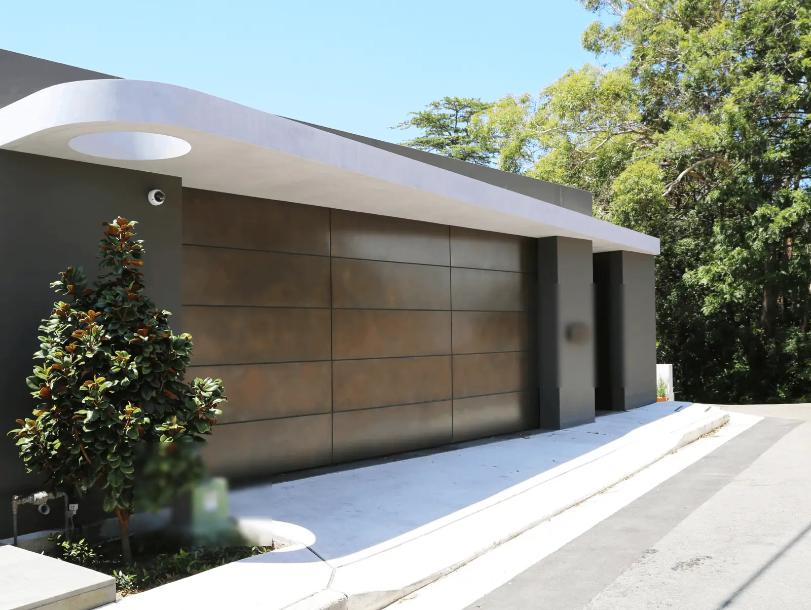 Double garage door in large flush panels with a brown bronze patina finish, under a curved white roof on a gray house.