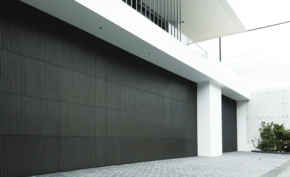 Two large charcoal garage doors with a fine vertical ribbed finish, set under the slab of a white modern house.