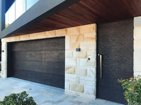 Double garage door in dark bronze rippled metal finish with no windows, beside a tall matching entry door in sandstone walls