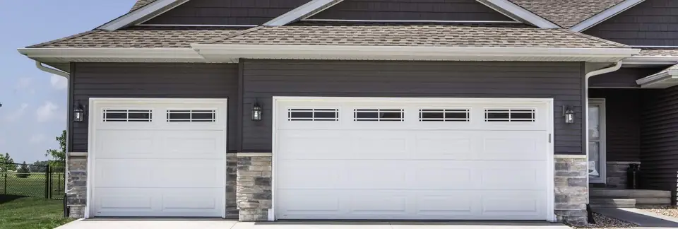 Two white long-panel steel garage doors, single and double, with prairie-grid windows across the top, on a gray-sided house with stone piers