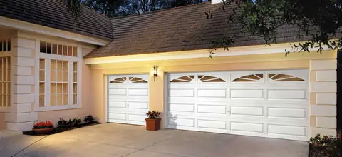 Two white long raised-panel steel garage doors with sunburst glass across the top, on a peach stucco house lit at dusk