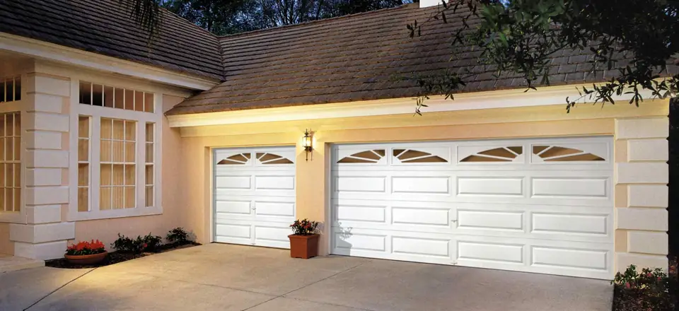 Two white long raised-panel steel garage doors with sunburst glass across the top, on a peach stucco house lit at dusk