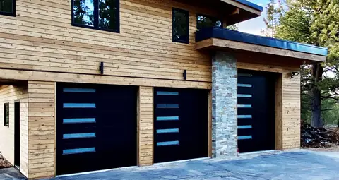 Three black flush steel single garage doors with slim glass windows stacked at the left, on a cedar-sided house