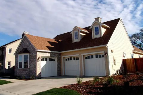 Three white single carriage-style steel garage doors with arched window tops, on a tan-sided house with a brick wing and two dormers