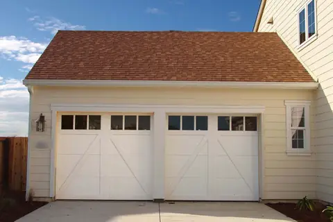 Two white single carriage-house garage doors with diagonal brace overlays and six square top windows each, on a tan-sided garage