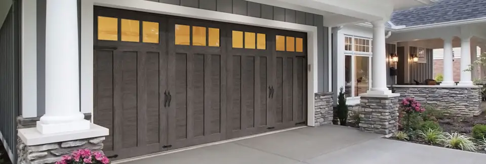 Weathered gray woodgrain carriage-house double garage door with lit square top windows and black handles, beside a porch with white columns