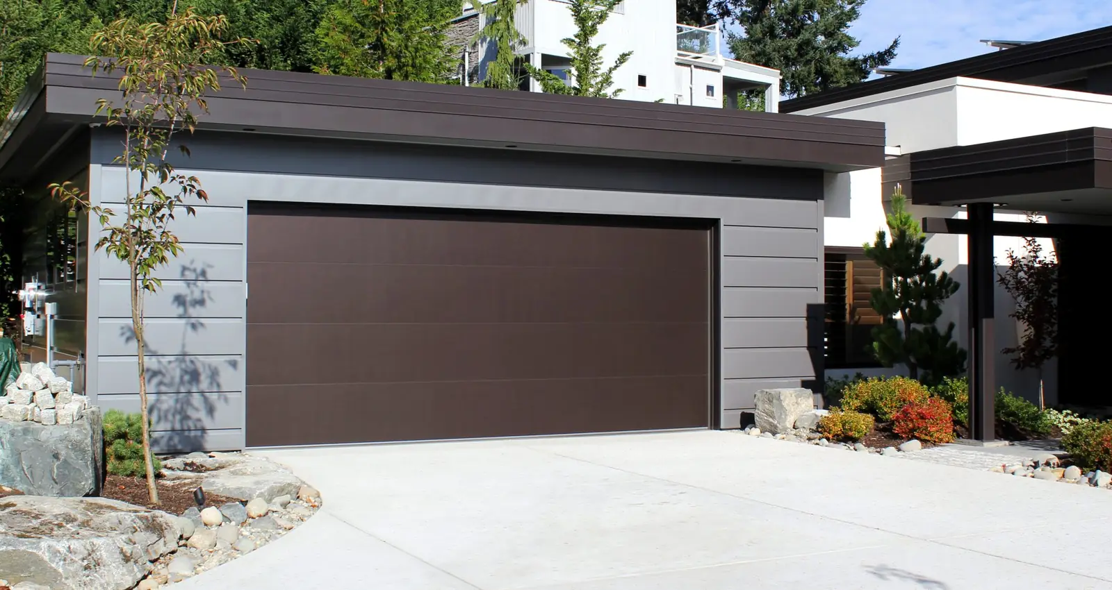 Flush double garage door in dark brown with no windows, on a flat-roof garage with gray horizontal siding and a concrete driveway.