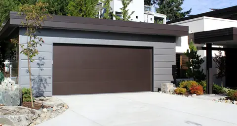 Flush double garage door in dark brown with no windows, on a flat-roof garage with gray horizontal siding and a concrete driveway.