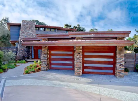 Two mahogany-stained wood garage doors, each with four long frosted glass bands, between stacked stone pillars under low copper-toned roofs.