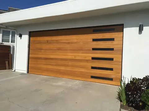 Double garage door of clear-finished cedar planks with six narrow black windows stacked on the right, on a white stucco house with a flat overhang.