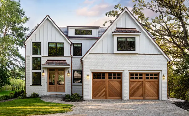 Two single cedar carriage-style garage doors with diagonal planks below and a row of square windows across the top, in a white painted-brick farmhouse.
