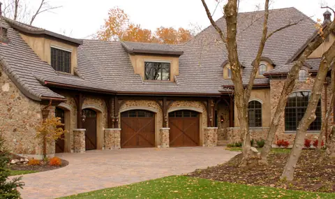 Two arched-top dark-stained wood carriage garage doors with X-brace panels and a row of windows under the arch, set in stone arches under timber brackets, beside two matching side doors.