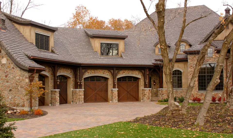 Two arched-top dark-stained wood carriage garage doors with X-brace panels and a row of windows under the arch, set in stone arches under timber brackets, beside two matching side doors.