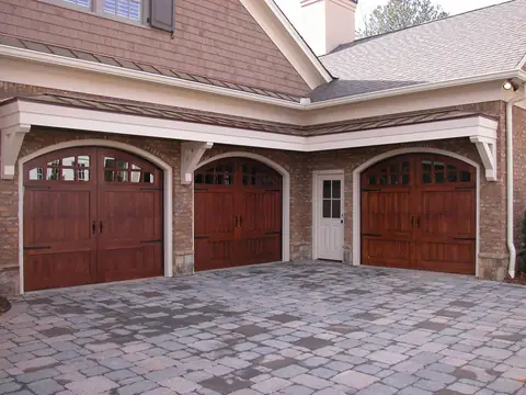 Three arched-top stained wood carriage garage doors with arched divided windows, black strap hinges and pulls, in brick arches around a white service door.