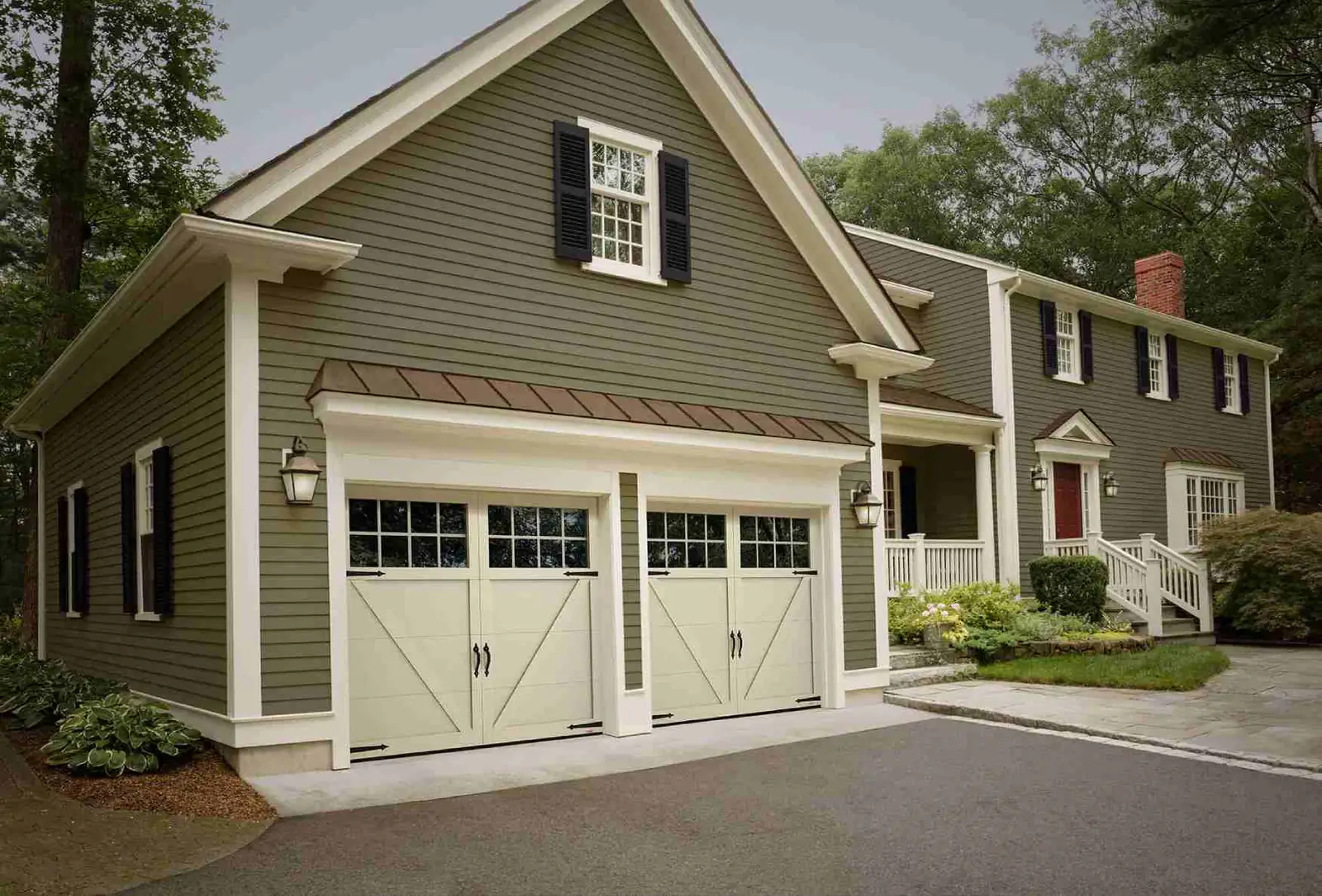 Two single almond carriage-style garage doors with V-brace panels, a top row of square windows and black strap hinges, on a green clapboard garage.