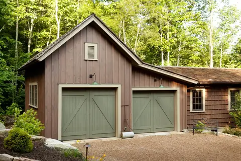 Two sage-green board-and-batten wood carriage garage doors with Z-brace panels and no windows, in a brown board-sided garage with a gravel court.