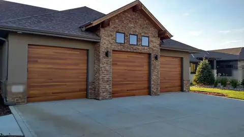 Three flush cedar plank garage doors with no windows, one in a taller stone-clad gable bay, on a stucco and stacked-stone house with a wide driveway.