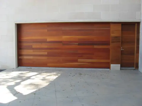 Flush double garage door of mixed-tone hardwood planks with a matching plank-clad side door, set in a pale limestone block wall.