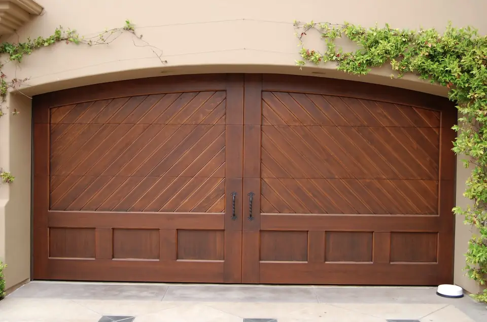 Arched-top double wood carriage garage door with diagonal planks above, recessed panels below and two twisted iron handles, in a tan stucco opening.