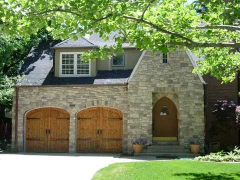 Two arched-top knotty wood carriage garage doors with clavos, black strap hinges and handles, in a stone house beside a pointed-arch front door.