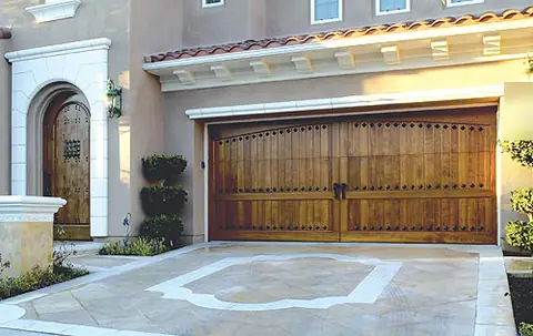 Double stained wood carriage garage door with rows of iron clavos and center handles, on a tan stucco house with a clay tile eave and inlaid concrete drive.