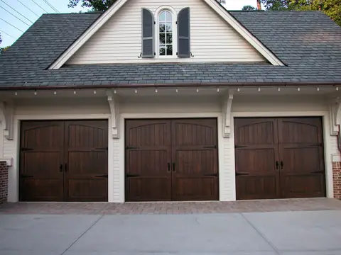 Three dark-stained wood carriage garage doors with arched top panels, black strap hinges and ring handles, on a cream clapboard garage with a dormer.
